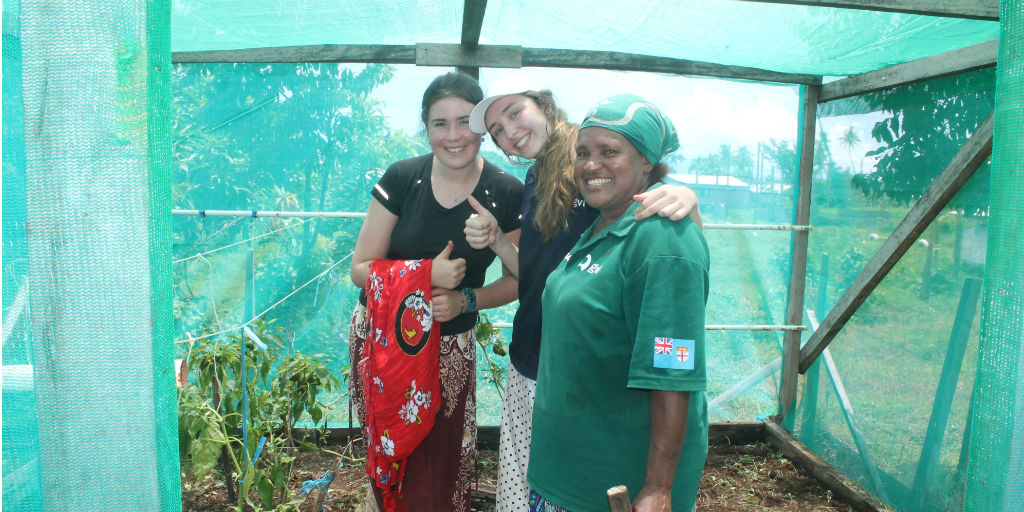 Two GVI volunteers standing next to a woman from Dawasamu in a plant shelter constructed out of wood and shade cloth.